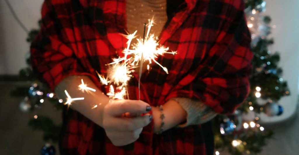 Close-up of hands holding sparklers in front of a lit Christmas tree.