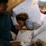 A father reading a bedtime story to his daughter, who is sleeping peacefully in bed.