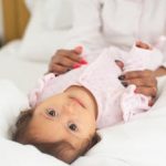 Charming baby in pajamas lying on white bed, attended by mother, creating a cozy morning scene.