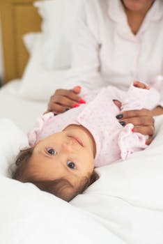 Charming baby in pajamas lying on white bed, attended by mother, creating a cozy morning scene.