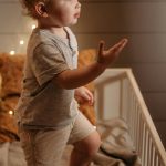 Cute toddler standing in a crib reaching out, surrounded by warm twinkling lights and cozy bedding.