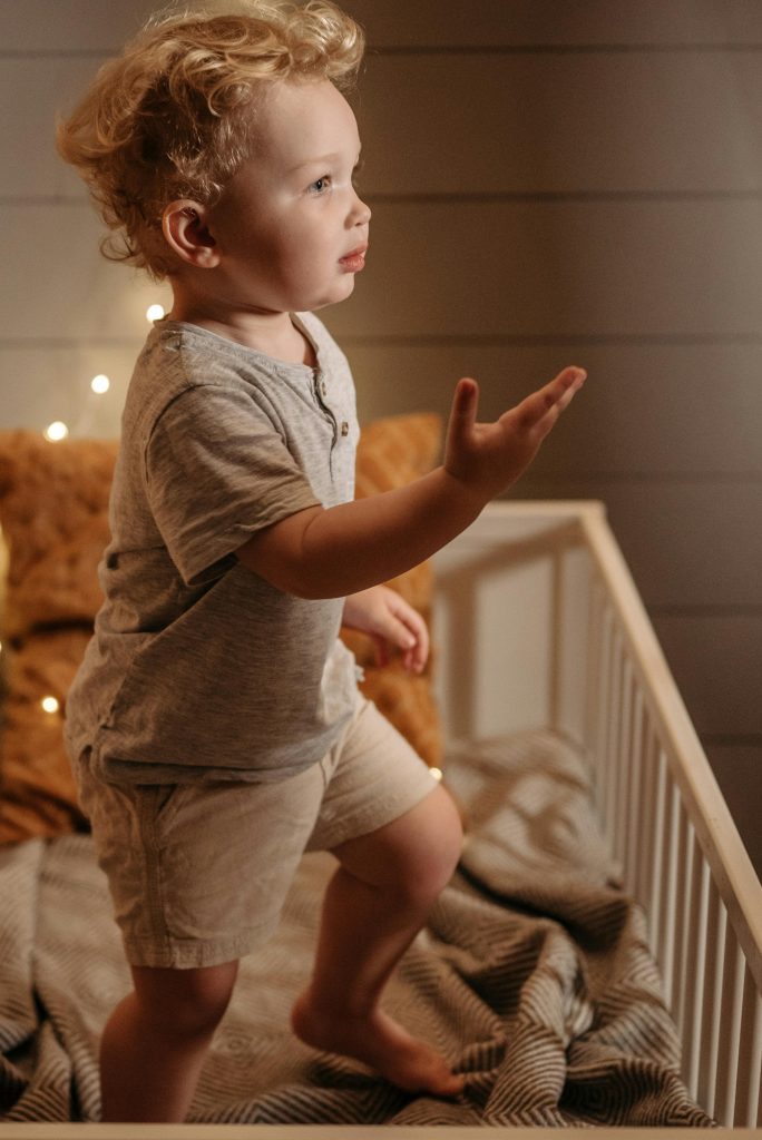 Cute toddler standing in a crib reaching out, surrounded by warm twinkling lights and cozy bedding.